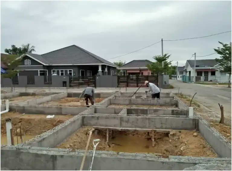 Kerja tapak rumah RumahHQ di peringkat asas — pekerja meratakan tanah di antara rasuk tanah konkrit yang sudah siap, di kawasan perumahan berjalan raya
