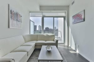 Modern apartment living room with white sectional sofa and city view through large windows.