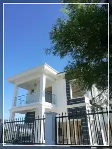 Modern white two-story house with balcony, black railing, and tree view against a clear blue sky.