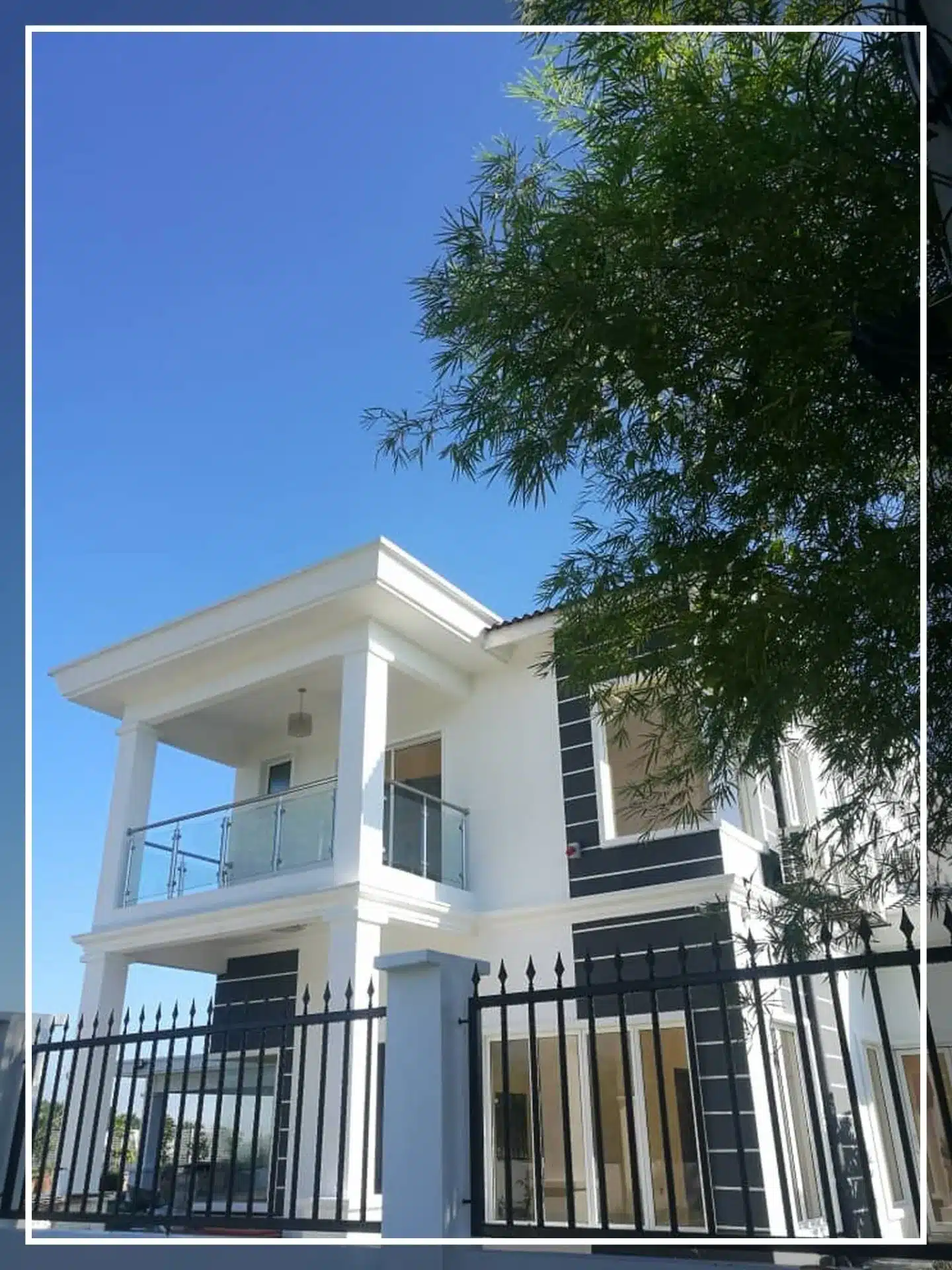 Modern white two-story house with balcony, black railing, and tree view against a clear blue sky.