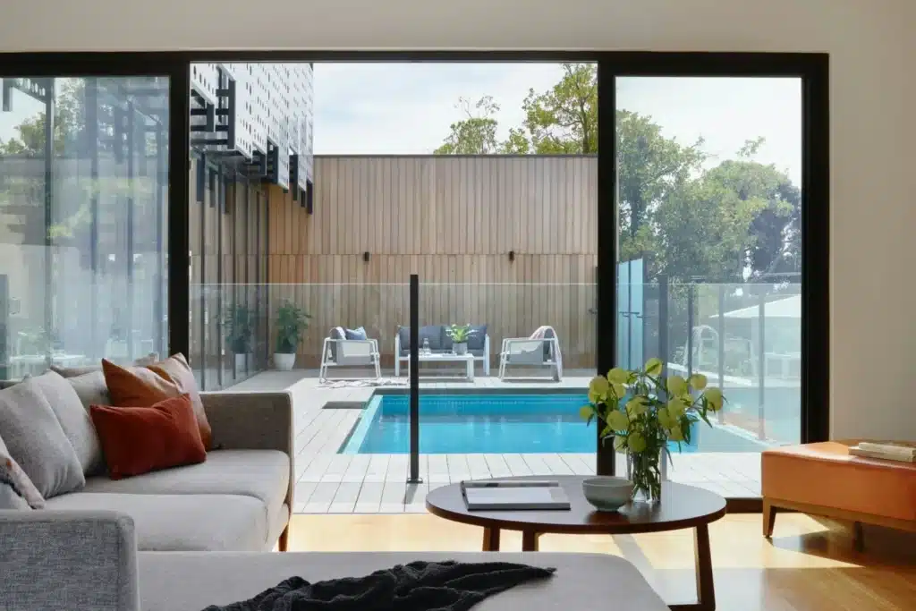 Modern living room with glass doors opening to patio and pool, featuring cozy furniture and decorative plants.