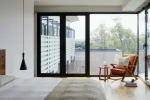 Modern bedroom with large windows, cozy chair, and wooden floors overlooking a lush view. Minimalist design.