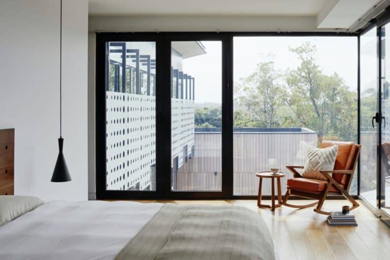Modern bedroom with large windows, wooden chair, and serene outdoor view. Minimalist decor, cozy and bright ambiance.