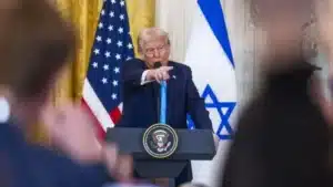 Speaker at podium with U.S. and Israeli flags in background, pointing towards audience during speech.