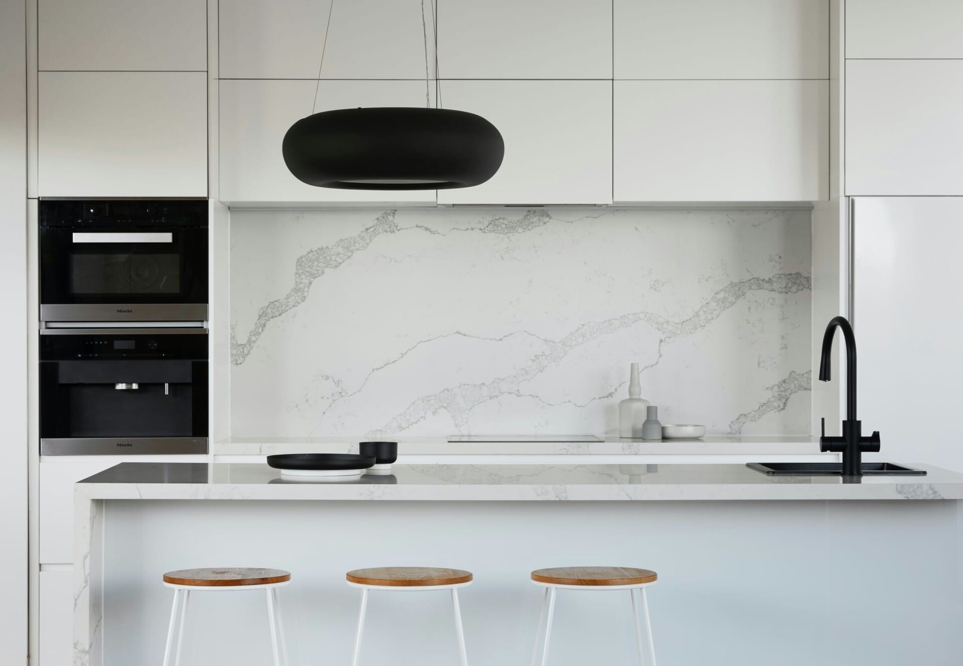 Minimalist white kitchen with marble backsplash, black faucet, modern pendant light, and wooden bar stools.