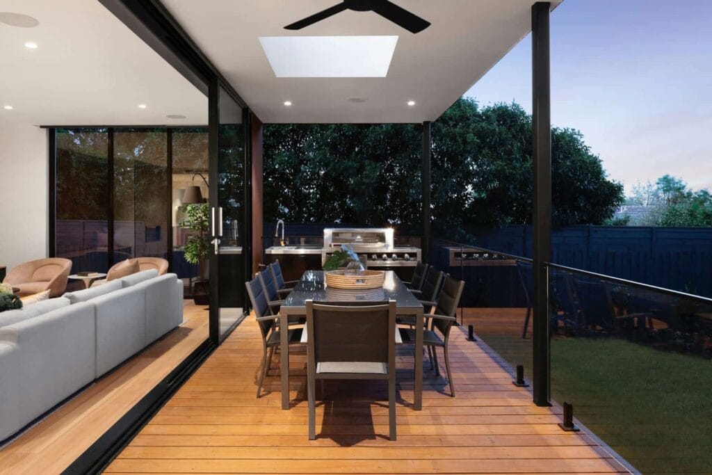 Modern patio dining area with outdoor kitchen, glass doors, and interior view of cozy living room.