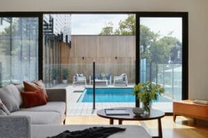 Modern living room view of outdoor pool with wooden deck and seating, through large glass doors.