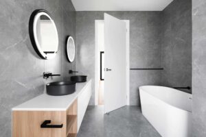 Modern bathroom with gray tiles, dual black sinks, round mirrors, and a white freestanding bathtub.