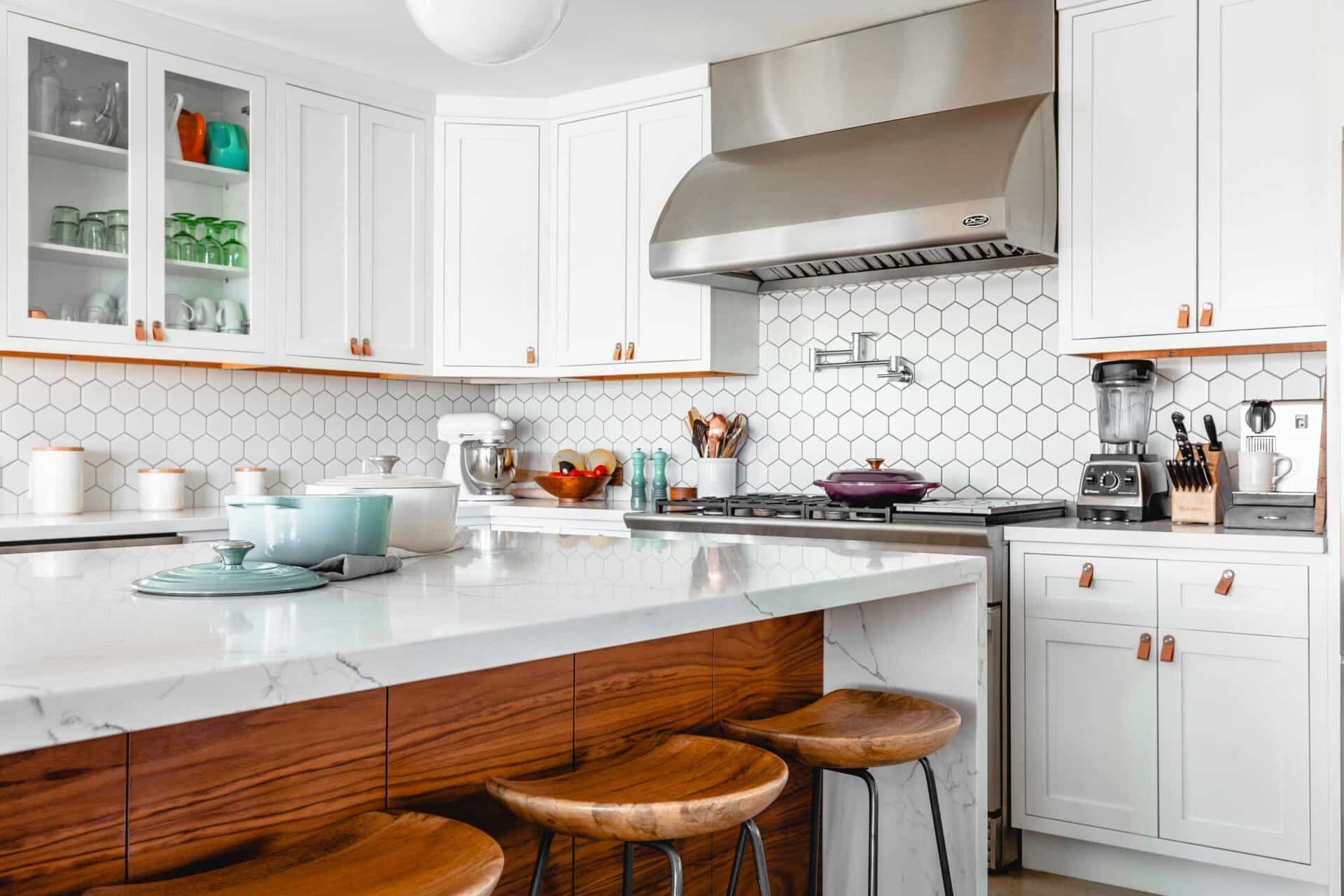 Modern kitchen with white cabinets, hexagonal backsplash, and wooden stools around a marble island counter.