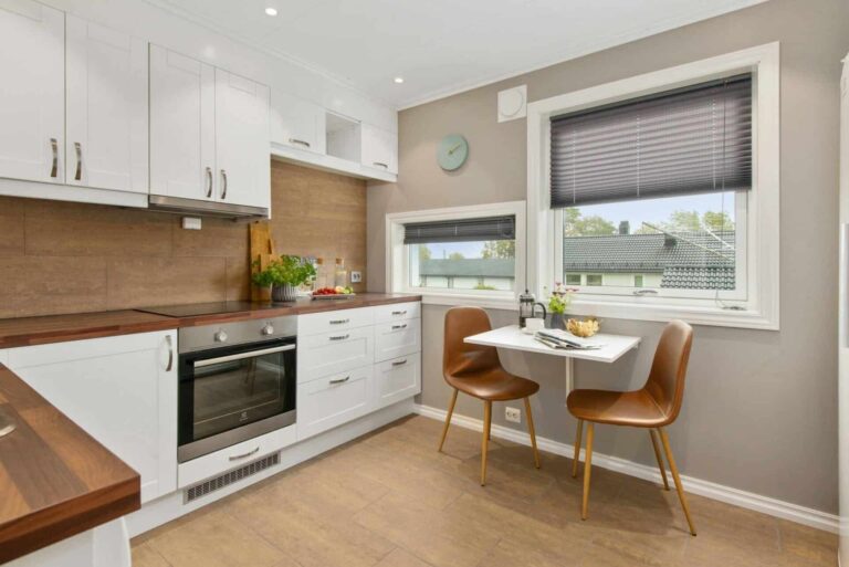 Modern kitchen with white cabinets, wood countertops, and a small dining table by a window with brown blinds.